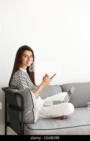 Photo verticale d'une femme indépendante qui a réussi à travailler à domicile sur un ordinateur portable. Fille assise sur un canapé avec un smartphone et un ordinateur, souriant à l'appareil photo, répondre Banque D'Images