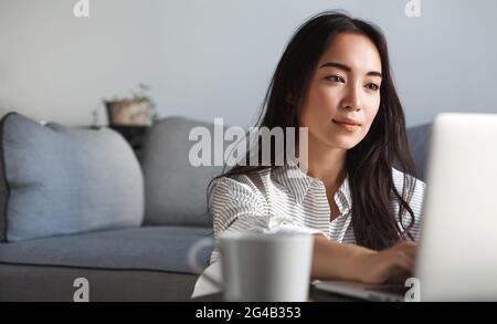 Femme d'affaires asiatique se reposant à la maison. Jeune fille étudiant e-learning sur ordinateur portable, travail assis dans la salle de séjour, boire du café et le shopping en ligne sur Banque D'Images