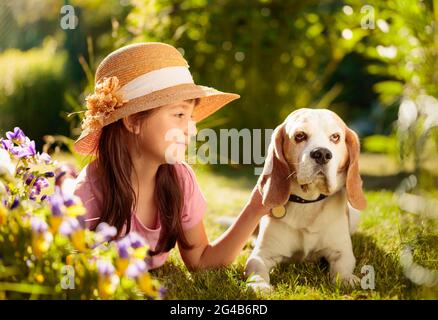 Une petite fille heureuse dans un chapeau de paille jouant avec son animal de compagnie sur l'herbe dans le jardin. Jour d'été ensoleillé dans le village. Banque D'Images