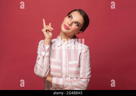 Une femme souriante contente vendeur pointe vers le haut, annonce l'article sur l'espace vide, a l'expression amicale du visage, porte une chemise décontractée sur fond rouge Banque D'Images