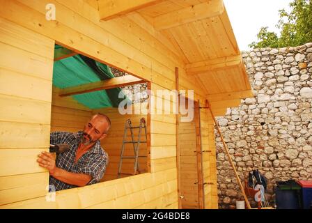 Un homme italien d'âge moyen construit une petite cabine préfabriquée en bois. Le toit est partiellement complet, la feuille de plastique vert est utilisée comme protecteur de pluie Banque D'Images