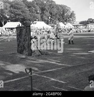 dans les années 1960, à l'extérieur, dans un champ, à un spectacle de comté, une exposition de moto, une photo montre un membre de l'équipe sur sa moto qui a effectué une cascade de daredevil dans une pile de bidons d'huile. À gauche, un obstacle construit en paille qui sera préparé, qui sera allumé, va dans les flammes et qu'un pilote d'affichage va aller, l'Angleterre, le Royaume-Uni. Les expositions acrobatiques et les cascades de motos étaient populaires à cette époque, avec les signaux royaux, les « casques blancs », une équipe d'exposition de l'armée britannique composée d'anciens pilotes de répartition, les plus connus. Ils avaient réalisé le premier affichage de cascades de moto enregistré en 1928. Banque D'Images