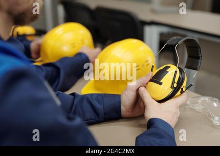 Gros plan d'un travailleur d'usine méconnaissable assis à une table avec un casque et des lunettes de protection et tenant un casque de protection insonorisant Banque D'Images