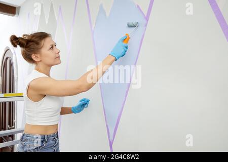 Femme souriante peignant un mur avec un rouleau de peinture et utilisant du ruban de masquage tout en se tenant sur l'échelle Banque D'Images