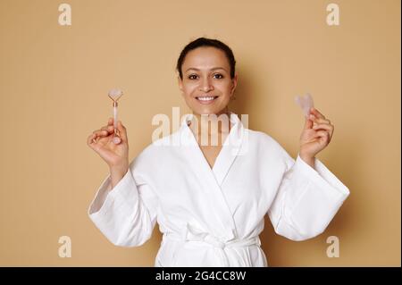 Femme d'âge moyen avec beau sourire en gaufre blanche peignoir posant avec gua sha et jade rouleau masseur dans ses mains isolées sur beige backgrou Banque D'Images