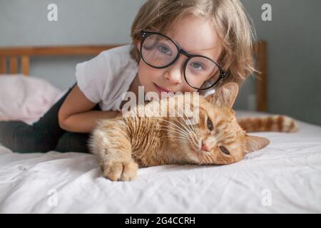 une petite fille avec des verres se trouve sur le lit et embrasse un gros chat de gingembre. Banque D'Images