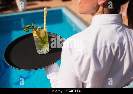 Une serveuse féminine sert un cocktail mojito sur un plateau au bord de la piscine. Une fête d'été folle Banque D'Images