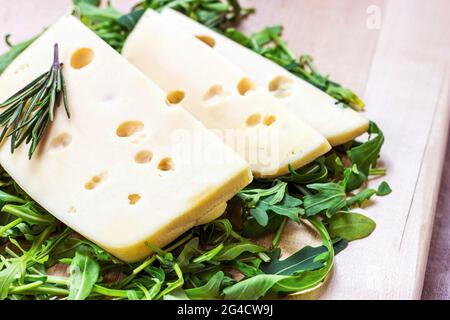 Tranches de fromage Maasdam avec feuilles d'arugula vertes sur fond de bois clair. Banque D'Images