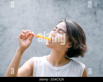 Femme mangeant des pop-sicles. Heureuse belle femme asiatique vêtue d'une chemise blanche sans manches décontractée tenant le pop-sicle jaune, à l'extérieur. Femme souriante qui m'apprécie Banque D'Images