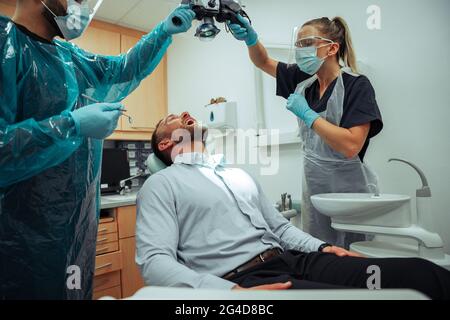 Client caucasien de sexe masculin assis sur une chaise de médecin ayant des dents examinées par des dentistes de sexe masculin et féminin Banque D'Images