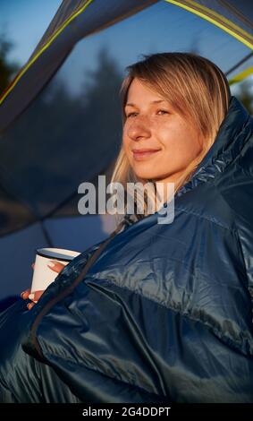 Gros plan d'une jeune fille enveloppée d'un sac de couchage assis dans une tente, tenant une tasse dans les mains et buvant du thé chaud le matin par temps ensoleillé à l'extérieur. Banque D'Images