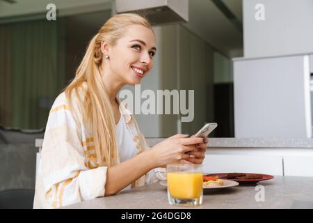 Femme excitée utilisant le téléphone mobile tout en prenant le petit déjeuner dans la cuisine à la maison Banque D'Images