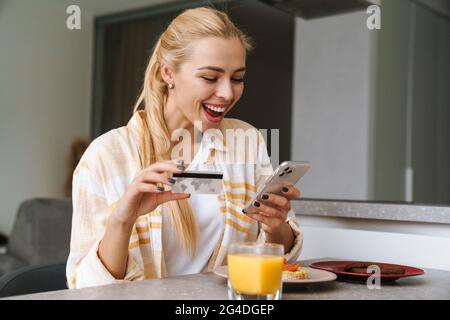 Femme excitée utilisant le téléphone cellulaire et la carte de crédit tout en prenant le petit déjeuner à la cuisine maison Banque D'Images