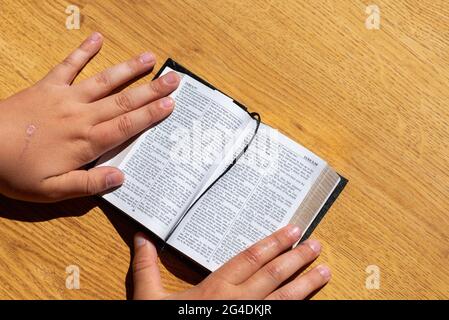 Les mains des enfants tiennent une bible penchée sur une table en bois. Petit livre Sainte Bible.Kid lecture de la sainte bible,closeup. Banque D'Images