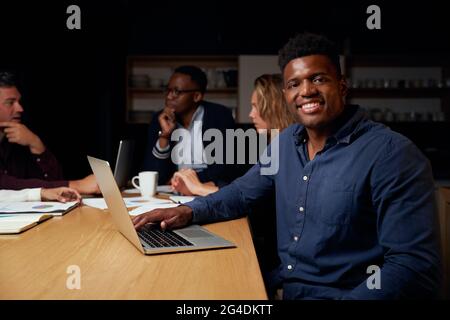Portrait d'un jeune homme africain souriant, chef d'équipe avec ordinateur portable, regardant l'appareil photo pendant que son collègue discute du projet en arrière-plan Banque D'Images