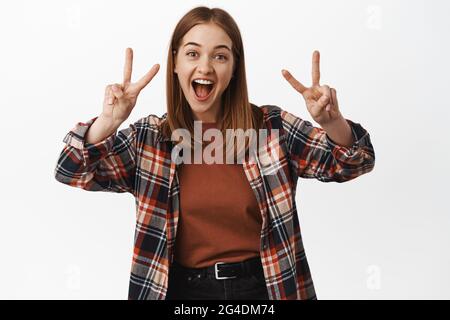 Positive et amicale jeune femme montre la paix V-signe geste, rire et sourire, posant heureux à l'appareil photo, debout dans des vêtements décontractés contre blanc Banque D'Images