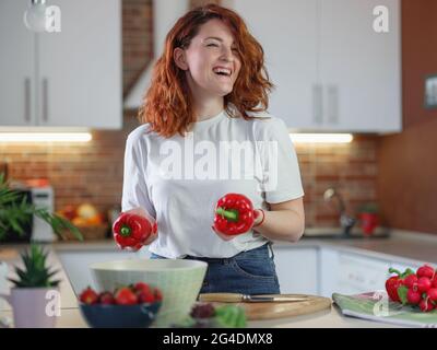 Belle redhair jeune femme prépare de la salade de légumes dans la cuisine. Alimentation végétalienne saine. Concept de régime. Cuisiner à la maison. Concept de blogueur de nourriture Banque D'Images