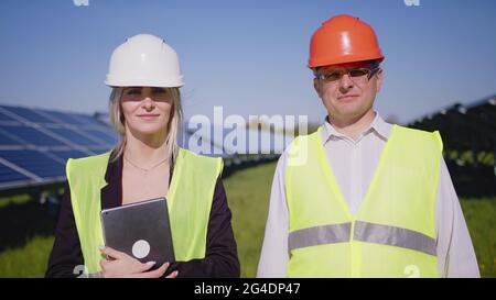 Portrait de deux ingénieurs en uniformes spéciaux avec casques de protection blancs. Belle femme et homme caucasien debout parmi les panneaux solaires et souriant Banque D'Images