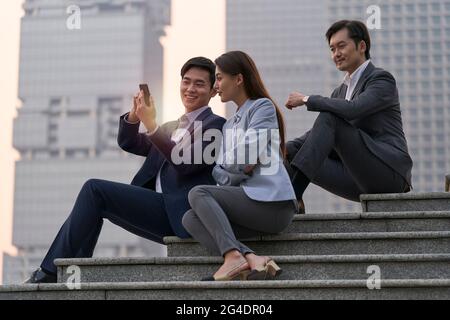 équipe de trois cadres d'entreprise asiatiques assis sur des marches regardant le téléphone mobile avec un fond de gratte-ciel Banque D'Images