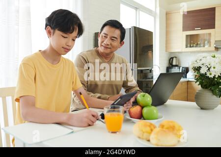 Père contrôlant son fils adolescent étudiant ou faisant des devoirs à la table de cuisine à la maison Banque D'Images