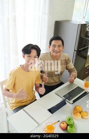 Joyeux adolescent et son père mûr assis à la table de kictehn avec une tablette et un ordinateur portable, montrant le pouce et souriant à l'appareil photo Banque D'Images