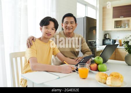 Portrait d'un père et d'un fils heureux qui s'embrasse assis à une table de cuisine lorsqu'ils travaillent et font leurs devoirs Banque D'Images