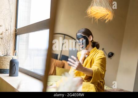 Femme en peignoir avec masque de nettoyage noir et téléphone dans la chambre Banque D'Images
