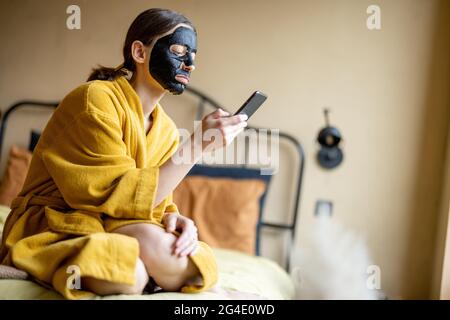 Femme en peignoir avec masque de nettoyage noir et téléphone dans la chambre Banque D'Images