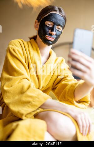 Femme en peignoir avec masque de nettoyage noir et téléphone dans la chambre Banque D'Images