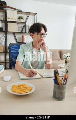 Un adolescent sérieux concentré dans des lunettes regardant un webinaire ou lisant un article sur ordinateur et écrivant dans un manuel à son bureau à la maison Banque D'Images