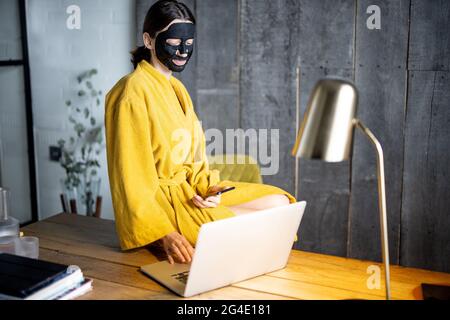 Femme en peignoir jaune avec masque de nettoyage noir sur son visage au bureau à domicile Banque D'Images