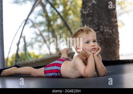 Portrait de mignon ennuyé petit caucasien drôle triste blond tout-petit garçon allongé à l'intérieur d'un grand trampoline noir à l'aire de jeux de la cour d'arrière-cour zone à l'extérieur chaude Banque D'Images