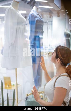 Femme caucasienne regardant une fenêtre de magasin et rêvant d'une robe. Banque D'Images