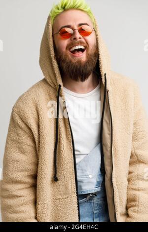 Beau jeune adulte modèle barbu homme portant une veste beige regardant l'appareil photo et riant à voix haute, exprimant positif et optimisme. Studio intérieur s Banque D'Images