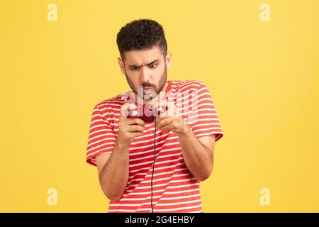 Addicted nerveux barbu homme dans rouge rayé t-shirt grimacing manette, jouer aux jeux vidéo en ligne, compétition virtuelle. Prise de vue en studio en intérieur Banque D'Images