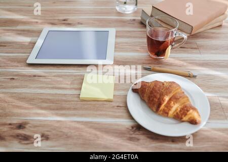 Tasse avec sac de thé et croissant sur l'assiette pour le petit déjeuner à côté de l'ordinateur tablette et pile de livres sur la table de la personne Banque D'Images