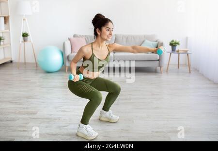 Bonne jeune femme indienne faisant des squats avec des haltères à la maison, pleine longueur Banque D'Images