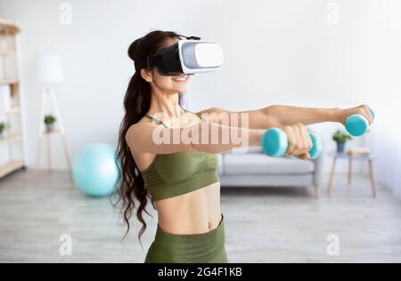 Concept d'entraînement virtuel. Mettre la jeune femme indienne dans des lunettes VR faire des exercices avec des haltères à la maison Banque D'Images