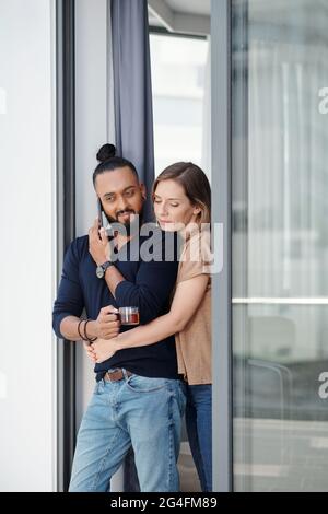 Jolie jeune femme qui embrasse son mari en train de boire du thé et de parler au téléphone avec un collègue ou un employé Banque D'Images