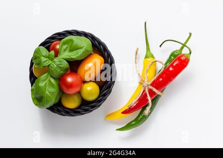 Tomates cerises dans un panier avec basilic et piment sur fond blanc. Vue de dessus des légumes frais. Banque D'Images