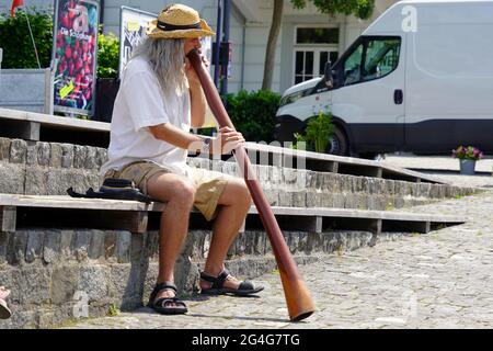 Homme jouant un didgeridoo dans la ville de Zug en Suisse. Il a de longs cheveux gris, porte un chapeau de paille, une chemise blanche, un short et des sandales. Banque D'Images