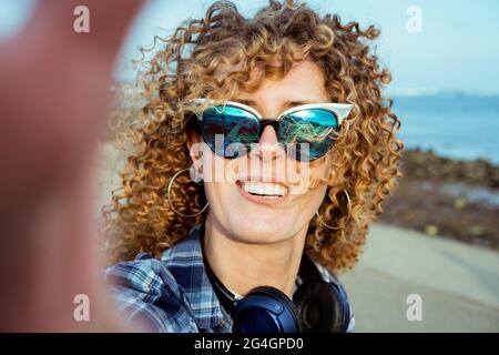 Gros plan sur les cheveux bouclés femme en lunettes de soleil faisant selfie photo sur l'appareil photo. Une jolie jeune femme avec casque prend le selfie tout en marchant près de la mer. Extérieur Banque D'Images