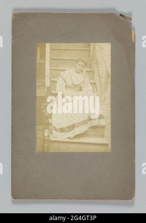 Photographie marron violet-rouge d'une femme afro-américaine non identifiée assise sur un escalier en bois. Elle est posée avec son bras gauche reposant sur son genou gauche. Sa tête repose sur sa main gauche. Sa main droite est placée sur ses genoux. Elle porte une robe de couleur claire avec une bande foncée autour de l'ourlet de la jupe. Les escaliers sont attachés à un bâtiment. La photo est collée sur un tapis gris foncé. L'arrière du tapis est vide Banque D'Images