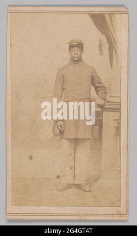 Une photographie brun rougeâtre d'un soldat non identifié de l'Union afro-américaine a posé dans un studio de photographe et#x2019; L'homme est légèrement décentré et porte un manteau de grenouille à neuf boutons avec un col haut. Le manteau présente des passepoils contrastés autour du col et des manches. Il porte également un pantalon droit, de lourdes bottes et un chapeau de képi à la coupe avec un bracelet en cuir. Il est debout, regardant directement la caméra, devant une rampe de colonnade avec des supports de roche cannelés. Bien qu'il soit trop fané pour voir, il y a probablement une ville ou un paysage idyllique derrière lui. Sa main gauche est soutenue Banque D'Images