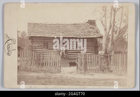 Photographie brun rougeâtre d'un groupe de familles afro-américaines en Géorgie. L'image montre une petite maison en rondins avec une clôture de piquet devant. La clôture est dotée d'une porte ouverte. Un arbre se trouve sur le côté droit de la photo. Derrière la porte se trouve la porte avant de la cabine avec trois marches menant à la porte. Les deux enfants sont assis sur la marche supérieure. Une fille portant une robe de couleur foncée est assise à gauche et un garçon portant des vêtements de couleur plus claire est assis à droite. Les deux enfants sont pieds nus. Une femme portant une robe de couleur claire avec un haut de couleur foncée et un foulard est debout behin Banque D'Images