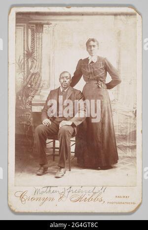 Une photo noir et blanc de carte de cabinet d'un homme et d'une femme afro-américains. L'homme est assis dans une chaise sur le côté gauche de la photo. Il porte un costume de couleur foncée et ses mains reposent sur ses genoux. La femme se trouve à côté de lui à droite. Elle porte une robe et une chemise de couleur foncée avec un mouchoir blanc attaché autour de son cou. Son bras droit est derrière l'homme tandis que sa main gauche est derrière son propre dos. La photo au bas de la carte est écrite sous “Mothers Friends!!” imprimée sous l'inscription au bas de la carte est “Cronyn & Banque D'Images