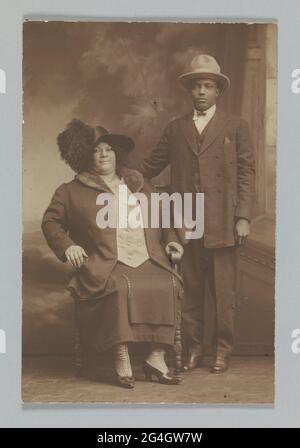 Une photographie brun violet-rouge profond des Afro-Américains, l'homme debout avec sa main sur le dos de la chaise. La femme porte un chapeau avec une grande plume. Les deux sujets font face à l'appareil photo. Le verso de la photo est vide. Banque D'Images