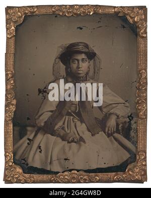 Cette photographie en noir et blanc est un portrait de trois quarts de longueur d'une jeune femme afro-américaine non identifiée assise avec son coude gauche reposant sur une table. La femme porte une robe rayée avec des épaules tombantes et des manches cloches qui ont une volants décoratifs le long de la couture extérieure et autour des poignets. Elle a sur un chapeau de paille de couleur foncée avec une couronne plate et un bord rond et plat avec une bande hachure à rayures. Un voile en filet noir avec dentelle fleurie décorative est fixé au chapeau et placé derrière le visage de la femme. Un large ruban de couleur foncée est attaché dans un arc autour du cou de la femme avec les extrémités exten Banque D'Images