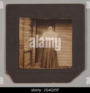 Photographie en noir et blanc d'une femme debout sur un port avec sa main droite sur la hanche. Elle porte une chemise et une longue jupe de style édouardien. La porte du bâtiment est derrière la femme et un numéro de maison [613] est visible au-dessus de la porte. Le timbre du photographe est apposé au dos de la photo. Banque D'Images