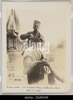 Soldats afro-américains servant en France pendant la première Guerre mondiale. Un soldat se rasse la tête d'un autre soldat. Un soldat est assis avec sa jambe droite traversée par-dessus sa jambe gauche. Ses mains reposent sur sa cuisse et il a une cigarette dans sa bouche. Son chapeau est suspendu sur un poteau en bois à sa droite. L'autre soldat se tient derrière. Ses mains sont levées et il tient la lame de rasoir dans cette main droite. Il porte son uniforme et son chapeau et il purge ses lèvres. Le logo signal corps USA se trouve dans le coin inférieur gauche de la photo. Banque D'Images